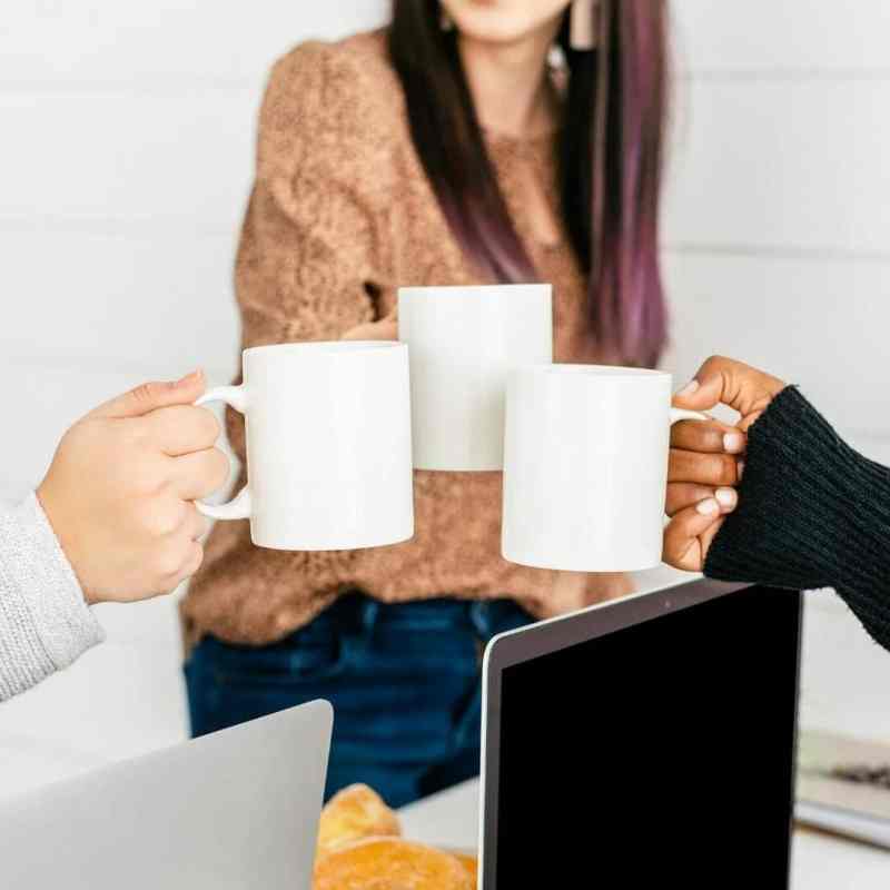 Three women clink coffee cups to in cheers. The women are not pictured. Instead, their hands are each holding a white mug over a table with laptops. 